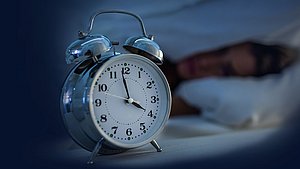 Selbsttest Schlafstörungen Ein Wecker zeigt kurz vor 4 Uhr nachts an, eine Person im Hintergrund liegt schlaflos im Bett.